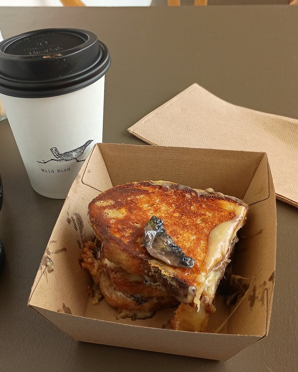 A mushroom toastie in a takeaway box next to a Wild Bird cup